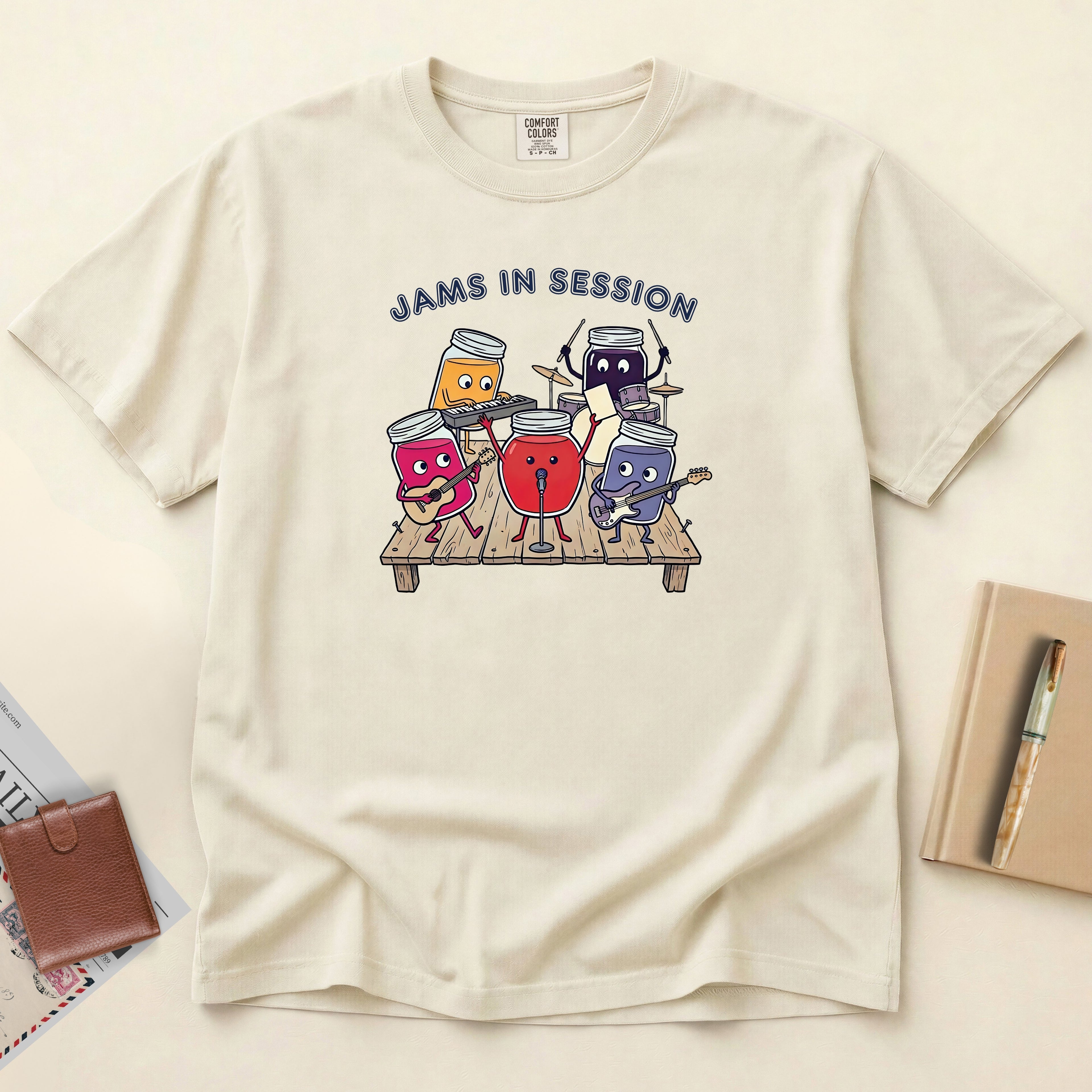 An off-white short-sleeve t-shirt with a colorful graphic of four cartoon drink cans playing musical instruments on a wooden stage, with the text 'JAMS IN SESSION' above them. The shirt is laid flat on a light beige background with a brown wallet, a book, and a pen nearby.